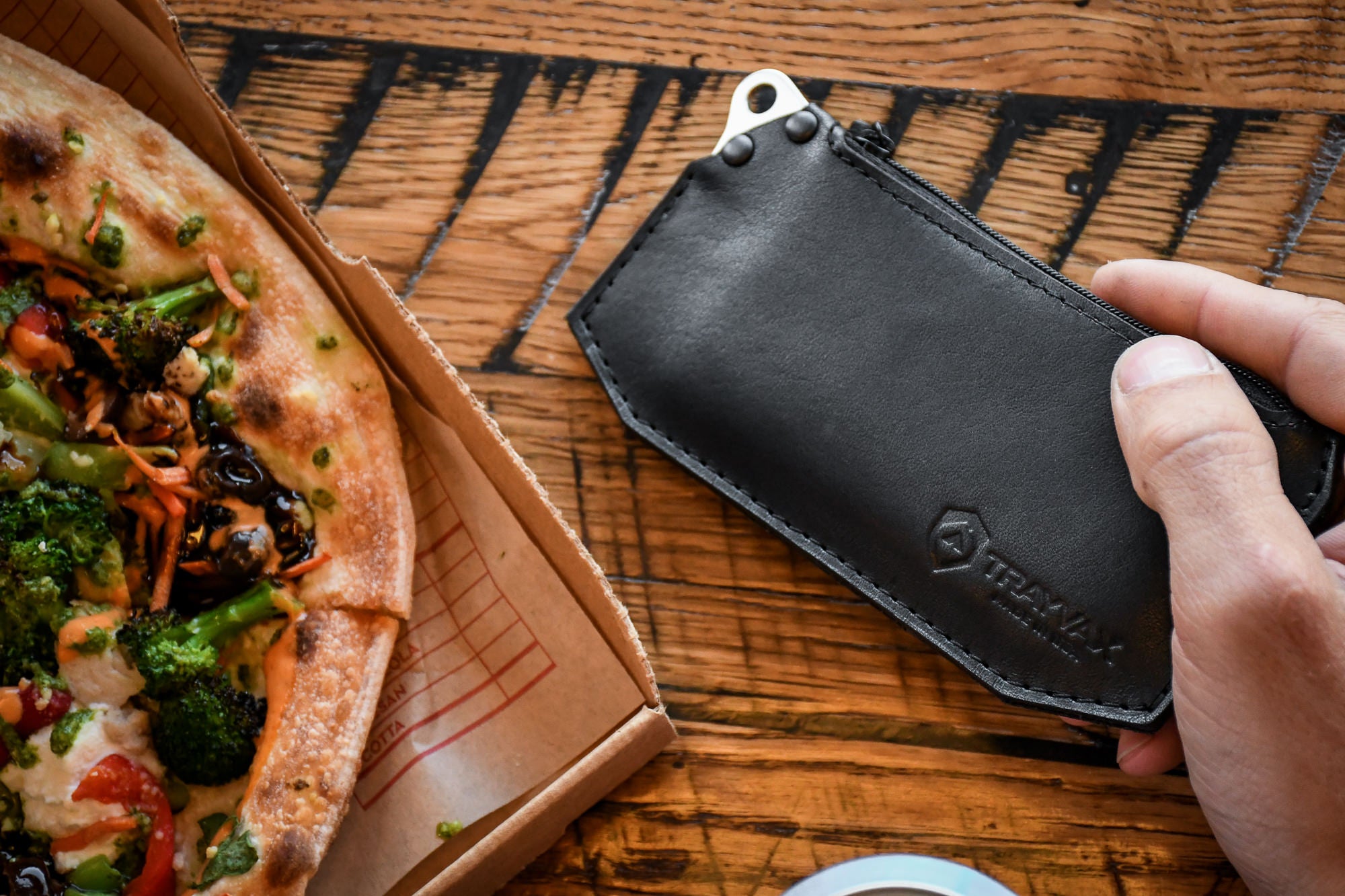 Man eating pizza on wooden table looking at his black leather Renegade Zipper Wallet